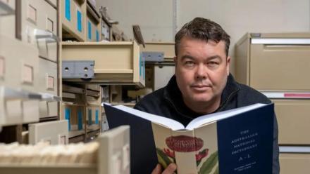Researcher Mark Gwynn holds open a copy of the Australian National Dictionary, surrounded by filing cabinets.