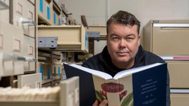 Researcher Mark Gwynn holds open a copy of the Australian National Dictionary, surrounded by filing cabinets.