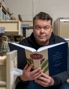 Researcher Mark Gwynn holds open a copy of the Australian National Dictionary, surrounded by filing cabinets.